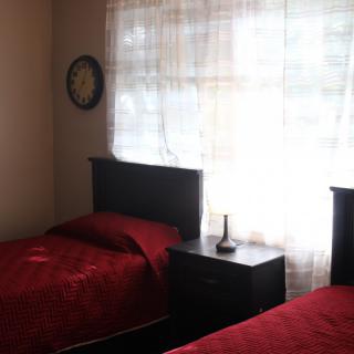 A cozy bedroom with two twin beds featuring red bedding, a window with sheer curtains, and a nightstand with a lamp.