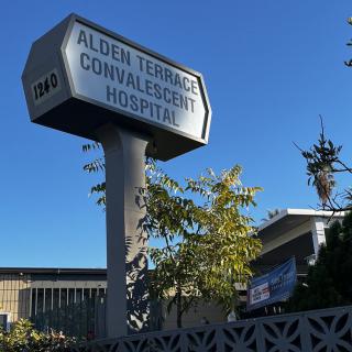 A sign for Alder Terrace Convalescent Hospital with surrounding trees and clear blue sky.
