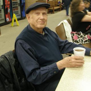 A man wearing a hat is sitting at a table holding a cup of coffee and smiling.