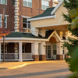 A cozy building entrance with a porch and brick exterior, surrounded by greenery.