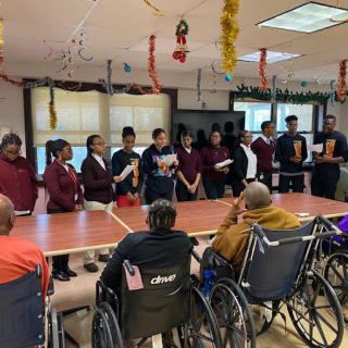 A group of people standing in a room with decorations, presenting to an audience seated in wheelchairs.
