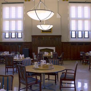 A spacious dining hall with round tables and chairs, featuring large windows and elegant chandeliers.