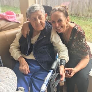 An elderly person sitting in a wheelchair with a younger woman hugging and smiling beside them outdoors.