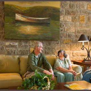 An elderly couple sitting on a sofa in a cozy living room with artwork and a stone wall.