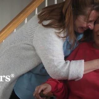 A woman lovingly hugs an older person seated at the bottom of a staircase, showing care and support.