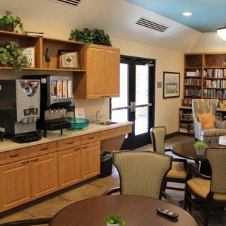 A cozy cafe area featuring a coffee station, tables, chairs, and a bookshelf.