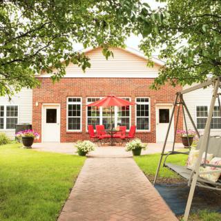 A cozy backyard with a brick house, featuring a patio set with red chairs and a swing, surrounded by lush green trees.