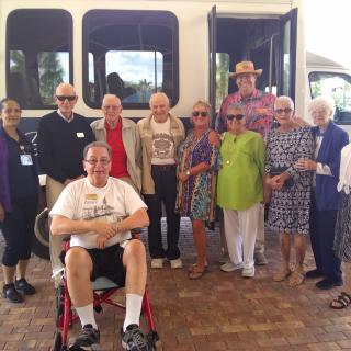 A group of seniors poses in front of a minibus, with one person seated in a wheelchair.