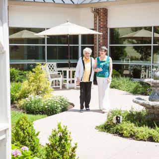 An elderly woman is walking with a caregiver in a sunny garden patio area.