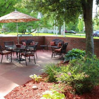 A serene patio area with tables and chairs under an umbrella, surrounded by lush greenery and well-maintained landscaping.