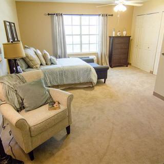 A cozy and well-furnished bedroom with a large window, bed, armchair, dresser, and neutral color scheme.