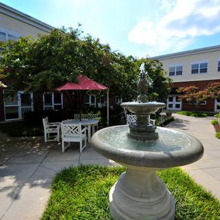 A tranquil courtyard featuring a fountain, outdoor seating with tables and chairs, shaded by umbrellas, surrounded by greenery and adjacent to a building.