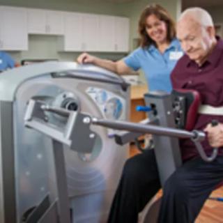 An elderly man receiving assistance from a therapist while using an exercise machine for rehabilitation.