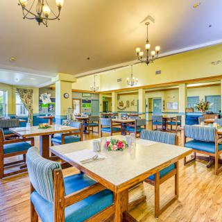 A brightly lit dining area with neatly arranged tables and chairs, featuring elegant chandeliers and wooden flooring.