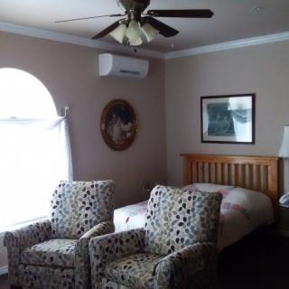 A cozy bedroom featuring two patterned chairs, a bed, ceiling fan, and wall-mounted air conditioner.