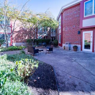 A charming outdoor patio area with seating, surrounded by greenery and adjacent to a red-brick building.