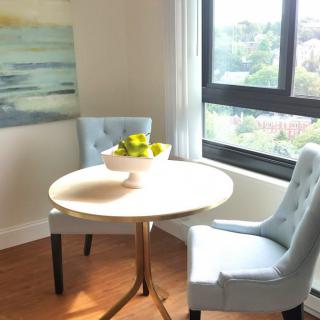 A cozy dining nook with a round table and two upholstered chairs, bathed in natural light from large windows.