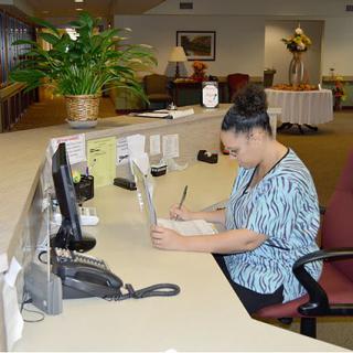 A receptionist working at a desk with a computer and writing notes in an office setting.