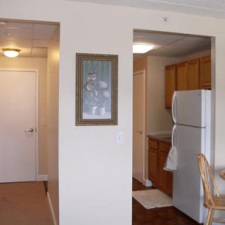 An interior view of an apartment showing a kitchen area with a refrigerator and wooden cabinets, adjacent to a hallway with a door.