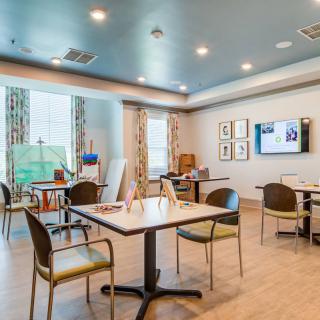 A modern, well-lit room with tables, chairs, and art supplies near large windows.