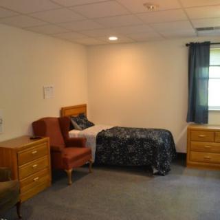 A simple bedroom with a single bed, armchairs, and wooden dressers.