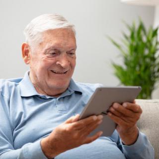 An elderly man smiling while using a tablet in his living room.
