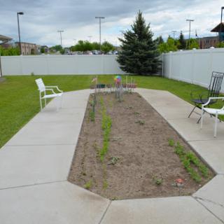 An empty garden area with a concrete pathway and chairs surrounding it, set against a backdrop of green grass and a cloudy sky.