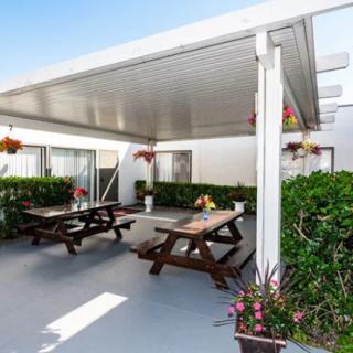 A cozy outdoor patio featuring picnic tables under a pergola, surrounded by greenery and decorated with hanging flower baskets.