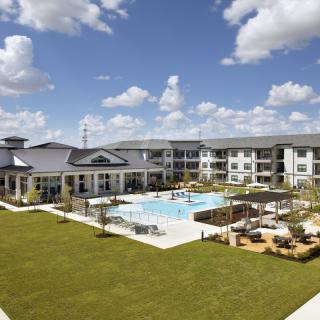 A modern apartment complex with a central swimming pool and a well-maintained lawn under a partly cloudy sky.