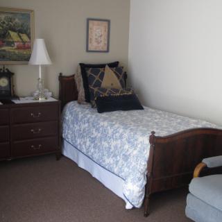 A cozy bedroom with a single bed adorned with patterned bedding, a wooden dresser with a lamp, and decorative art on the walls.
