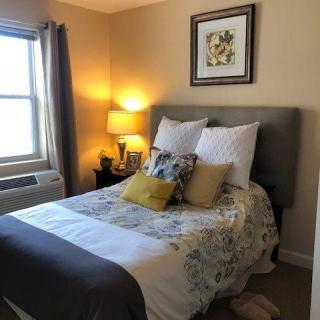 A cozy bedroom with a neatly made bed, decorative pillows, a bedside lamp, and a window providing natural light.