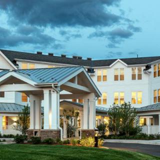 A large residential building with a well-lit entrance and landscaped surroundings under a cloudy evening sky.