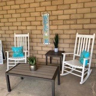A cozy patio setup with white rocking chairs, decorative pillows, and plants against a brick wall.