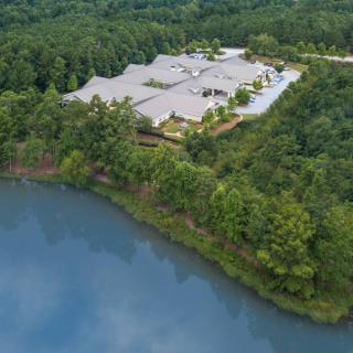 Aerial view of a large building surrounded by dense forest and adjacent to a tranquil lake.