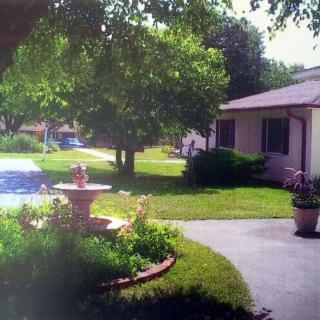 A peaceful garden scene with a fountain, surrounded by lush greenery and a house in the background.