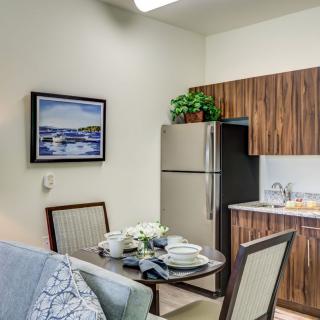 A small kitchen and dining area with a table set for two, featuring a refrigerator, cabinets, artwork on the wall, and decorative plants.