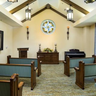 A peaceful chapel interior featuring wooden pews, an altar with candles and flowers, a stained glass window, and a piano.