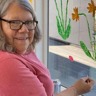 A woman wearing glasses is painting colorful flowers on a window.