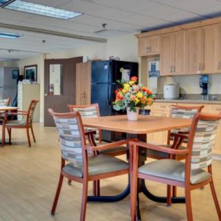 A well-lit dining area with tables and chairs, featuring a kitchen in the background and a vase of flowers on the table.