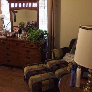 A cozy bedroom corner with a patterned armchair, wooden dresser with mirror, and various decorations.