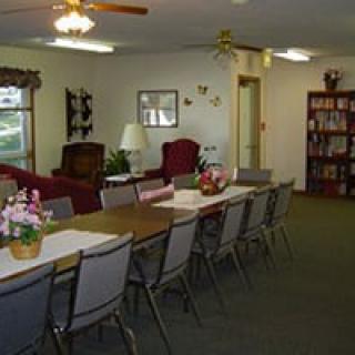 A communal dining or meeting area with long tables and chairs, featuring a bookshelf and ceiling fans.