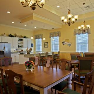 A spacious dining area with a wooden table and a kitchen in the background, featuring elegant chandeliers and large windows.