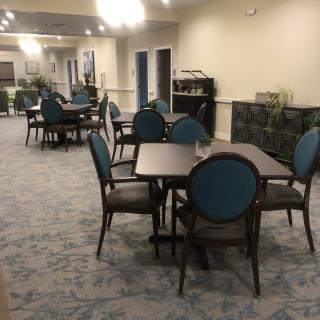 An indoor dining area with multiple tables and chairs arranged on a carpeted floor, complemented by overhead lighting and decorative plants.
