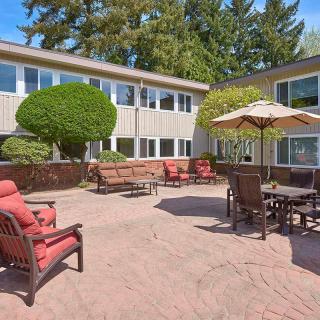 A spacious outdoor patio area featuring comfortable seating and tables under umbrellas, surrounded by a building with large windows and trees in the background.