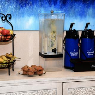 A refreshment station featuring a beverage dispenser, a fruit basket, water bottles, and muffins.