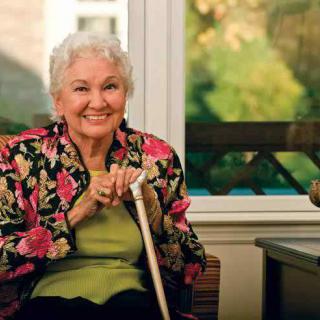 An elderly woman with a bright smile sits indoors, holding a cane and wearing a floral jacket.