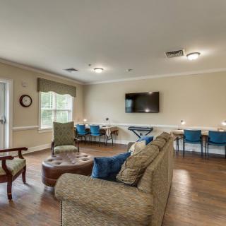 A spacious living room featuring comfortable seating, a TV on the wall, and a row of chairs along the back wall.