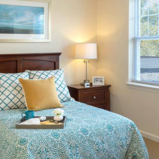 A cozy bedroom with a neatly made bed, featuring patterned bedding and a decorative pillow. A lamp sits on the nightstand next to the window.