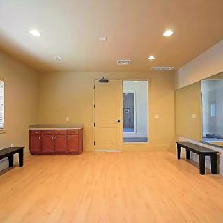 A spacious, empty room featuring wooden flooring, benches, and natural light pouring in from the windows.