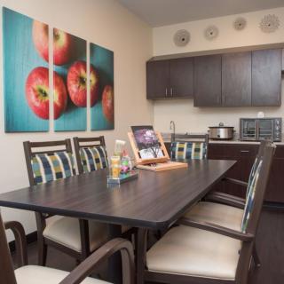 A modern dining area with a table, cushioned chairs, vibrant artwork of tomatoes, and kitchen appliances in the background.